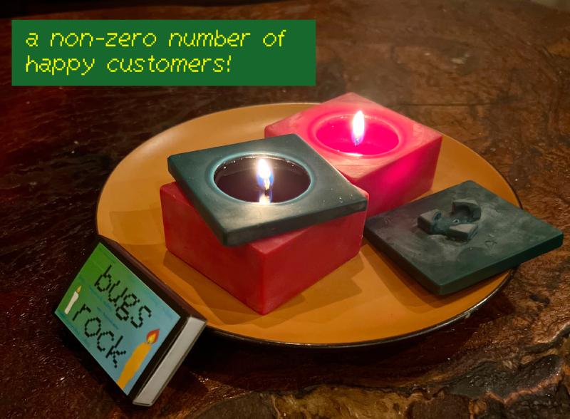 photo of my "nice tomato" candles, both lit but one lit without the green top on it. rested nearby are a box of bugs rocks matches. this is staged on a nice reclaimed wood table that my best friend lisa owns. i added the caption "a non-zero number of happy customers!"
