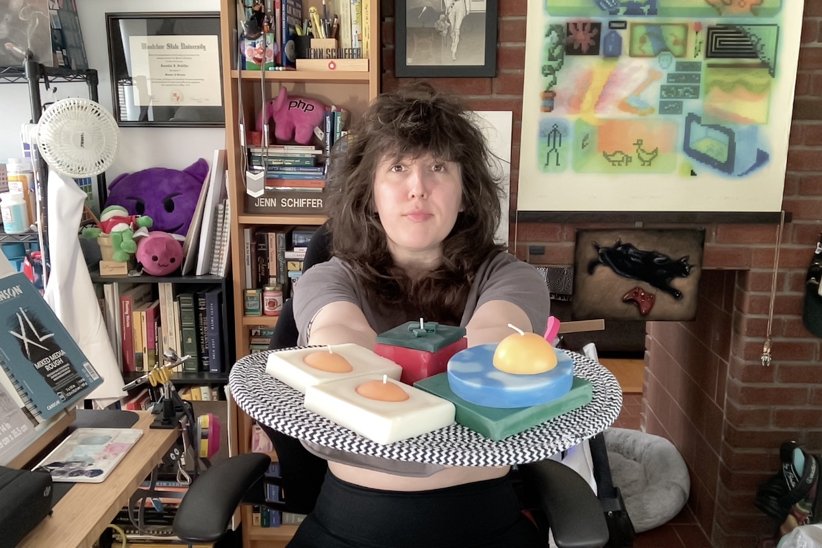 photo of me in my home office surrounded by art and gizmos and doo-dads. i am holding up a mat to the camera and on it are my three first collection candles: a duo of eggs, a tomato, and an outdoor grass/sky/sun sculpture"